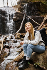 Smiling woman sitting with bottle of water and looking away in forest near waterfall