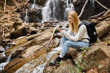 Naklejka premium Calm blonde young woman with phone near mountain waterfall in the forest while hiking