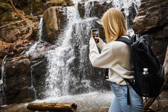 Back view of blonde young woman taking photo of mountain waterfall in the forest while hiking