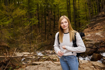 Naklejka na ściany i meble Pretty blonde female hiker wearing sweater and jeans crossing the forest creek walking in woods