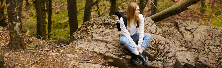 Naklejka na ściany i meble Young woman hiker with backpack sitting at halt on rocky cliff in forest looking away, banner