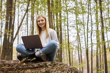 Deurstickers Smiling bright woman on halt in woods, sitting on boulder with laptop in lotus position © LIGHTFIELD STUDIOS