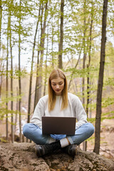 blond relaxed woman on halt in woods, sitting on boulder with laptop in lotus position