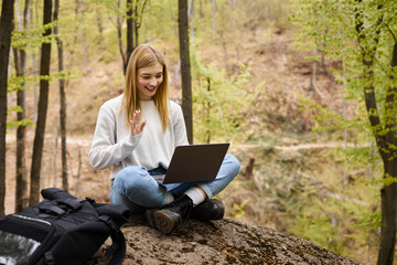 Young blonde woman with backpack in woods, sitting on boulder with laptop and making video calls