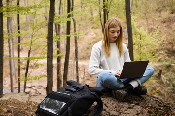 Young blonde woman with backpack hiking in woods, sitting on boulder with laptop and working