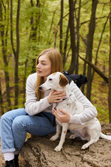 Caring blonde woman hugging her dog, while having a halt on forest trip, both looking away