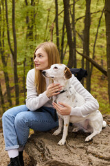 Smiling blonde woman hugging her dog, while having a halt on forest trip, both looking away