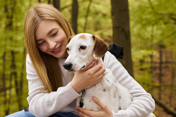 Smiling blonde woman hugging her dog, caring for her pet while having a halt on forest trip