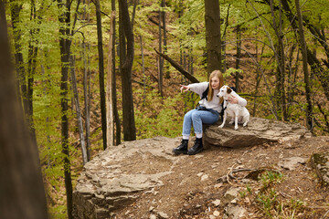 Devoted blond adventurer showing her curious dog a direction sitting on rock in the woods