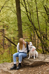 Loving blonde hiker, dressed in jeans and cozy, sweater, petting her loyal dog sitting in forest
