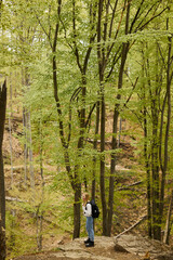 Top view of blonde woman adventurer in cozy sweater and jeans wearing backpack, standing in forest