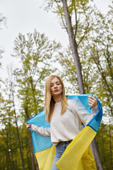 Calm blonde woman outdoors in forest covering body with Ukrainian national flag as cape