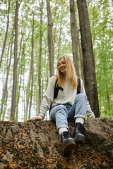 Naklejka na ściany i meble Smiling hiking blonde woman wearing sweater and backpack sitting on a rock in woods