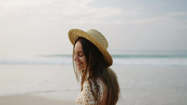Charming girl in flowery dress posing, smiling at seaside, foamy water waves. Young brunette woman in straw hat walking at ocean shore. Stylish female tourist flirting, going relaxed at the seashore.