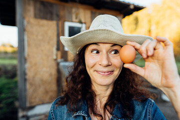 Portrait of funny joyful farmer woman holding fresh egg near her face. While enjoying farming, a female farmer holds a fresh egg Happy moment of collecting eggs
