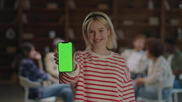 A confident young woman stands in front of a therapy group, holding out a smartphone with a green screen for a digital presentation
