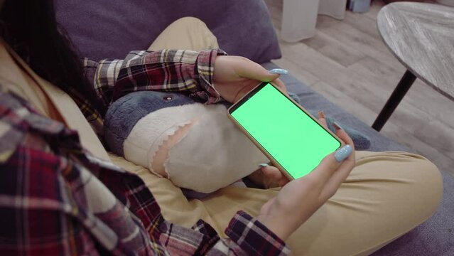 young woman using smartphone with green screen mockup in home living room to watch videos while lying on sofa, High quality 4K footage