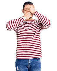 Fototapeta premium Young handsome man wearing striped sweater covering eyes and mouth with hands, surprised and shocked. hiding emotion