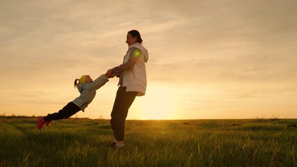 Mom circles her little daughter at sunset in spring. Happy family is playing in park. Kid, daughter playing with mom, baby flying, mom holding hands. Child dreams of flying. Mother and kid silhouette