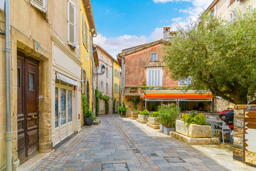 A small square leading to a narrow alley full of shops and sidewalk cafes in the historic old town of the hilltop medieval village of Grimaud, France in Provence.