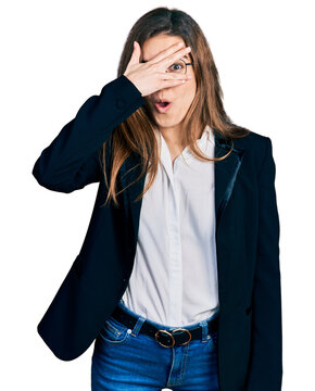 Young caucasian girl wearing business style and glasses peeking in shock covering face and eyes with hand, looking through fingers with embarrassed expression.