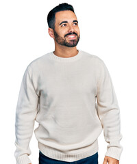 Young hispanic man with beard wearing casual white sweater looking away to side with smile on face, natural expression. laughing confident.