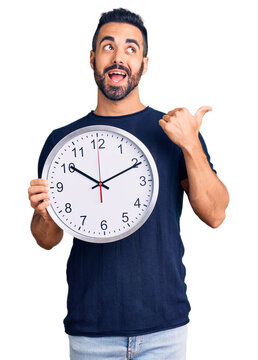 Young hispanic man holding big clock pointing thumb up to the side smiling happy with open mouth