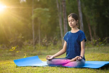 Fotobehang peaceful young indian girl sitting in a padmasana yoga position or lotus pose with her eyes closed in the forest at a sunrise time © Dharma