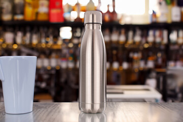 One metal vacuum-insulated bottle for water or coffee and a cup of coffee on a table on a bar background. - obrazy, fototapety, plakaty