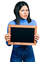 Young brunette woman with blue eyes holding blackboard clueless and confused expression. doubt concept.