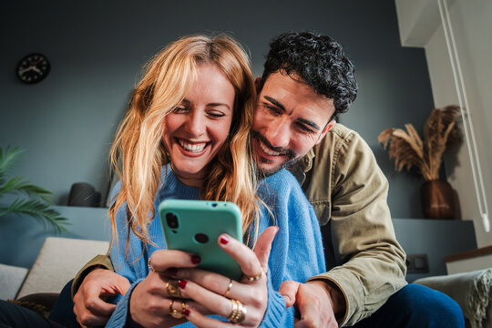Young caucasian couple smiling using a cellphone to do shopping online. Man and woman watching the social media with a smartphone. Adult friends having fun browsing on internet with a mobile phone - Powered by Adobe