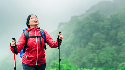 Banner with adult Asian woman enjoying a walk on a mountain trek, light spring rain and fog in the mountains, holding Nordic walking poles, active senior lifestyle