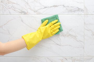 crop of female hand in yellow rubber glove cleaning a white marble ceramic tiles on the wall in the...