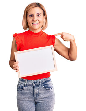 Young blonde woman holding empty white chalkboard pointing finger to one self smiling happy and proud
