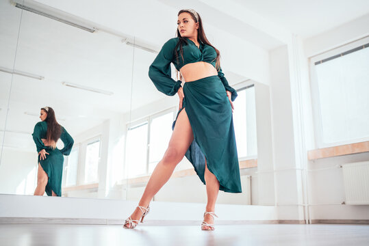 Graceful woman dressed dark green Latin dancing dress, high heels shoes doing elegant dance pas in white color big hall with big mirror wall. People's expressions, beauty of woman's body concept image