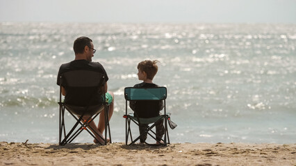 Father and son in chairs on beach in front of wild sea waves, admiring the nature and enjoying the...