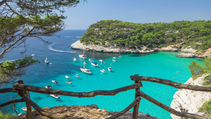 Yachts on turquoise Mediterranean sea cove surrounded by green pines woods seen from Cala Mitjana cliff panoramic viewpoint in Menorca Island, Spain