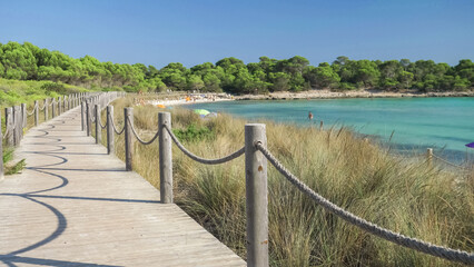 Wooden path along Son Saura sandy beach with turquoise water surrounded by green pine trees in...