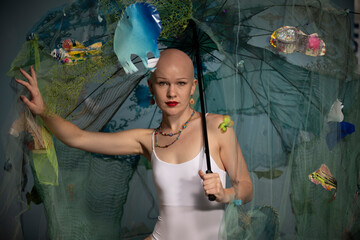 A bald woman in white attire holding an umbrella decorated with ocean life elements against a blue backdrop