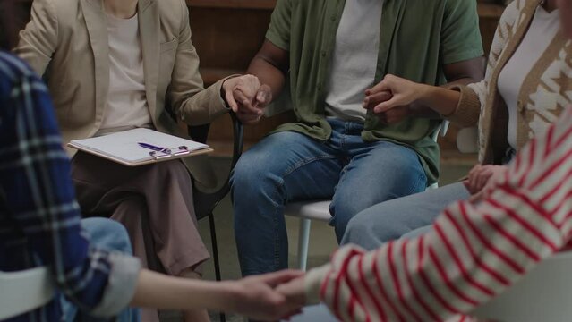 A group of diverse individuals participate in a supportive handholding exercise during a group therapy session