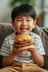 Excited happy asian little boy eating big burger at home