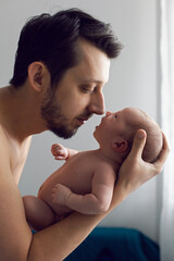 Close-up fashionable portrait of a man with hipster beard without outerwear holding a baby girl in his arms standing in front of a window.