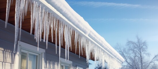 Long, sharp icicles are hanging from the edge of a house roof. The sun is shining, causing the icicles to glisten in the light.