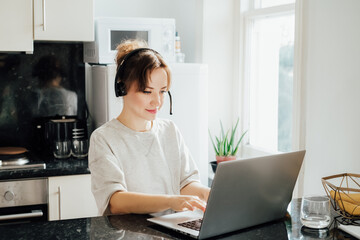 Obraz na płótnie Canvas Remote work at home. Woman in headset working on Laptop at Kitchen Table in tiny flat. Student has online classes. Manager has video meeting. Customer support operator in telemarketing service