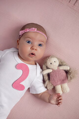 girl baby infant with a bow lies on the bed in a white bodysuit with a doll toy next to it, the child has a pink number 2 months on a pink background
