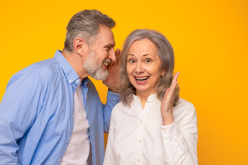 Cheerful senior husband sharing his secret with wife, yellow backdrop