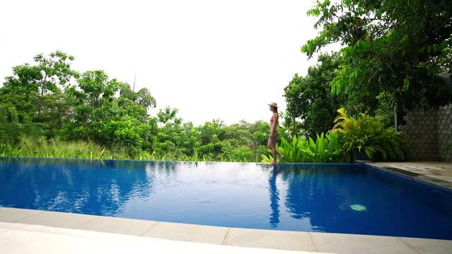 Woman walks by infinity pool in luxury villa, tropical green plants background. Gentle stroll, enjoys lush scenery, peaceful mood. Leisure travel, exclusive destination, relaxation in nature. Slowmo