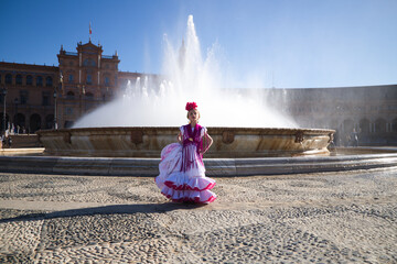 A pretty little girl dancing flamenco dressed in a white dress with pink frills and fringes in a famous square in seville, spain. The girl has a flower in her hair. In the background a big fountain. - obrazy, fototapety, plakaty