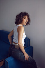sexy fashionable young curly-haired woman in jeans and white T-shirt sitting on the blue sofa against the white wall of the house. showing white pants.