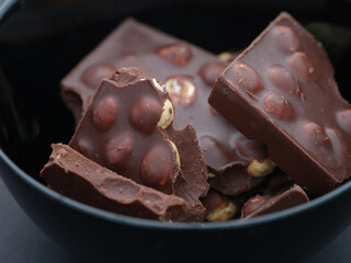Broken pieces of dark chocolate with whole hazelnuts in a black bowl. Close up.